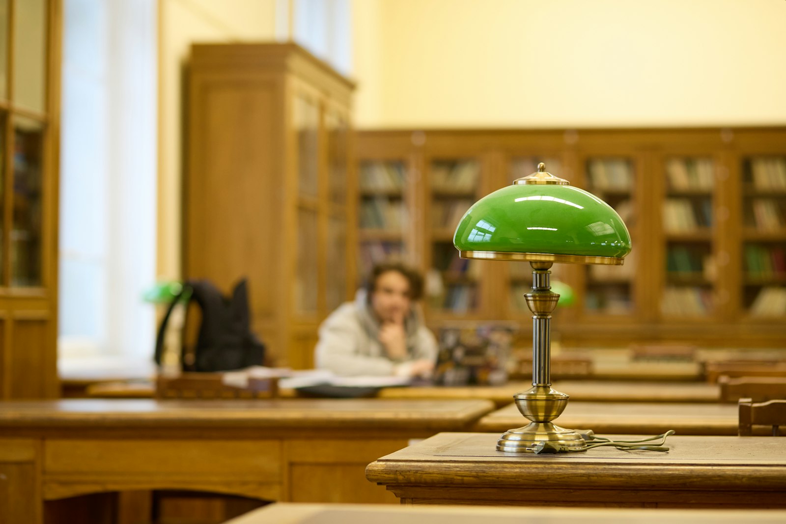 A reading room with a green desk lamp, long tables and shelves of books